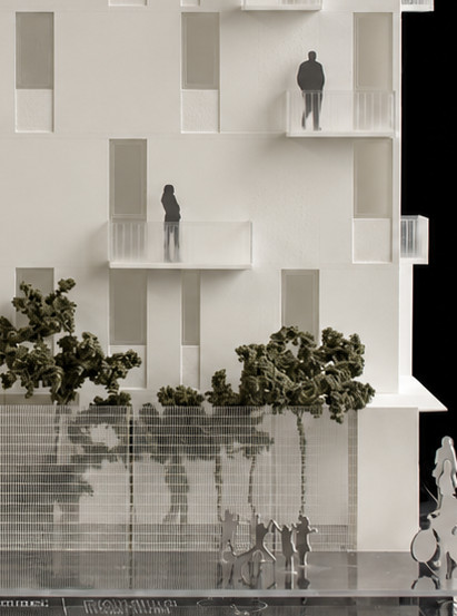 Maqueta frontal de un edificio blanco de cuatro niveles con balcones, vanos verticales, árboles y figuras a escala sobre una base de acrílico, fotografiada sobre fondo oscuro.